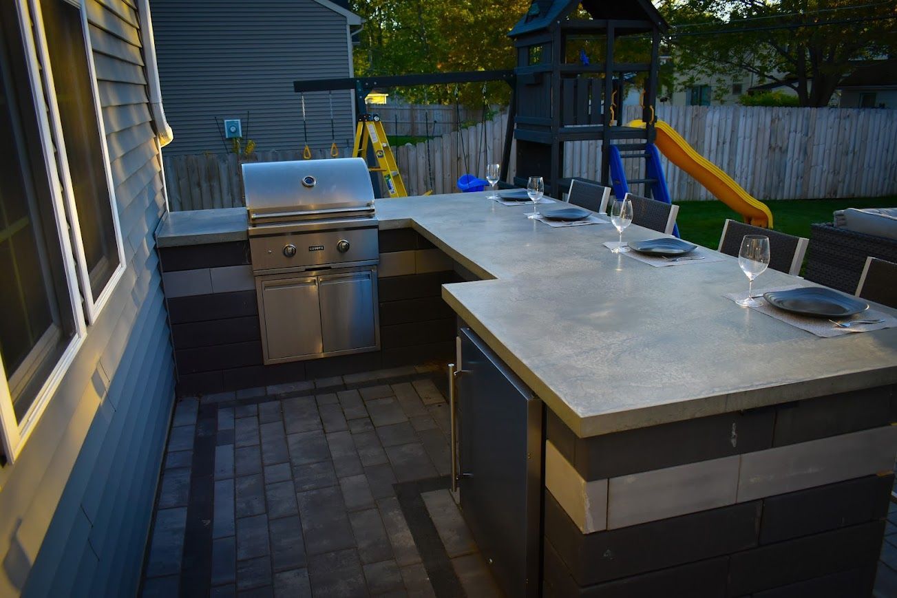 Outdoor kitchen with grill, countertop, and dining area on a paved patio. Includes playground in the background.