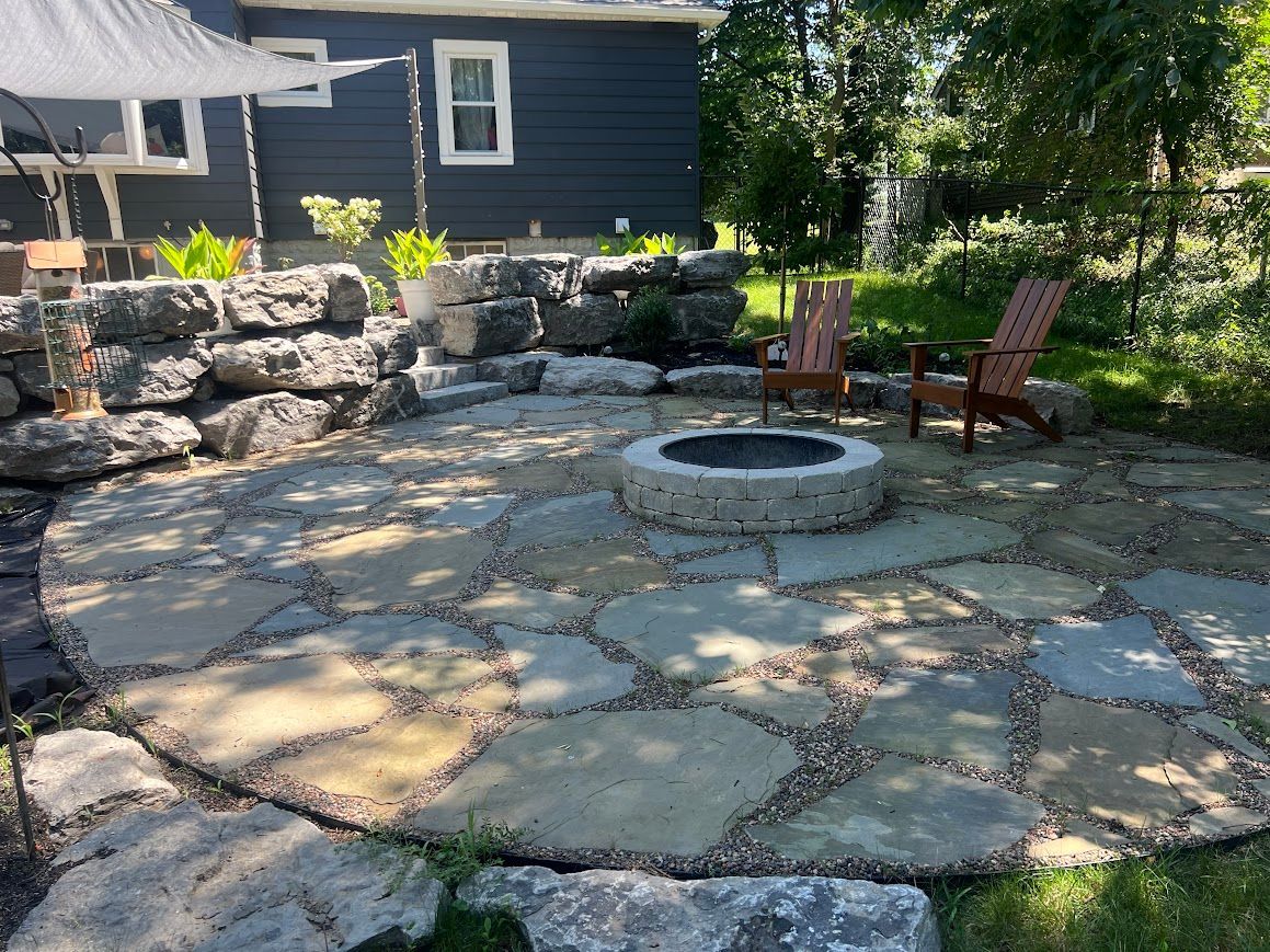 Stone patio with a fire pit, two wooden chairs, and a dark blue house in the background.