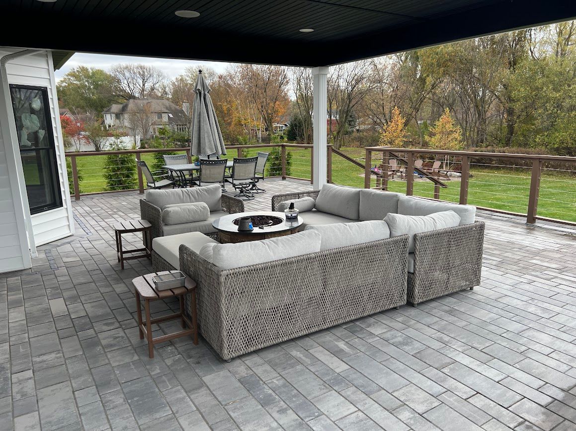 Gray wicker sectional sofa on a paved patio, with side tables and a fire pit, overlooking a grassy yard.