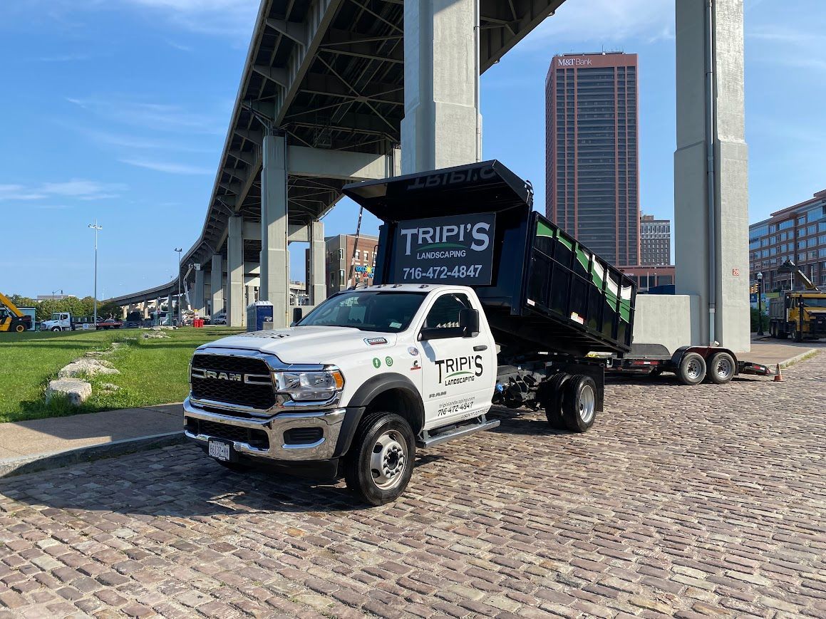 White Tripi's dump truck parked under a bridge. Buildings and construction equipment visible in the background.