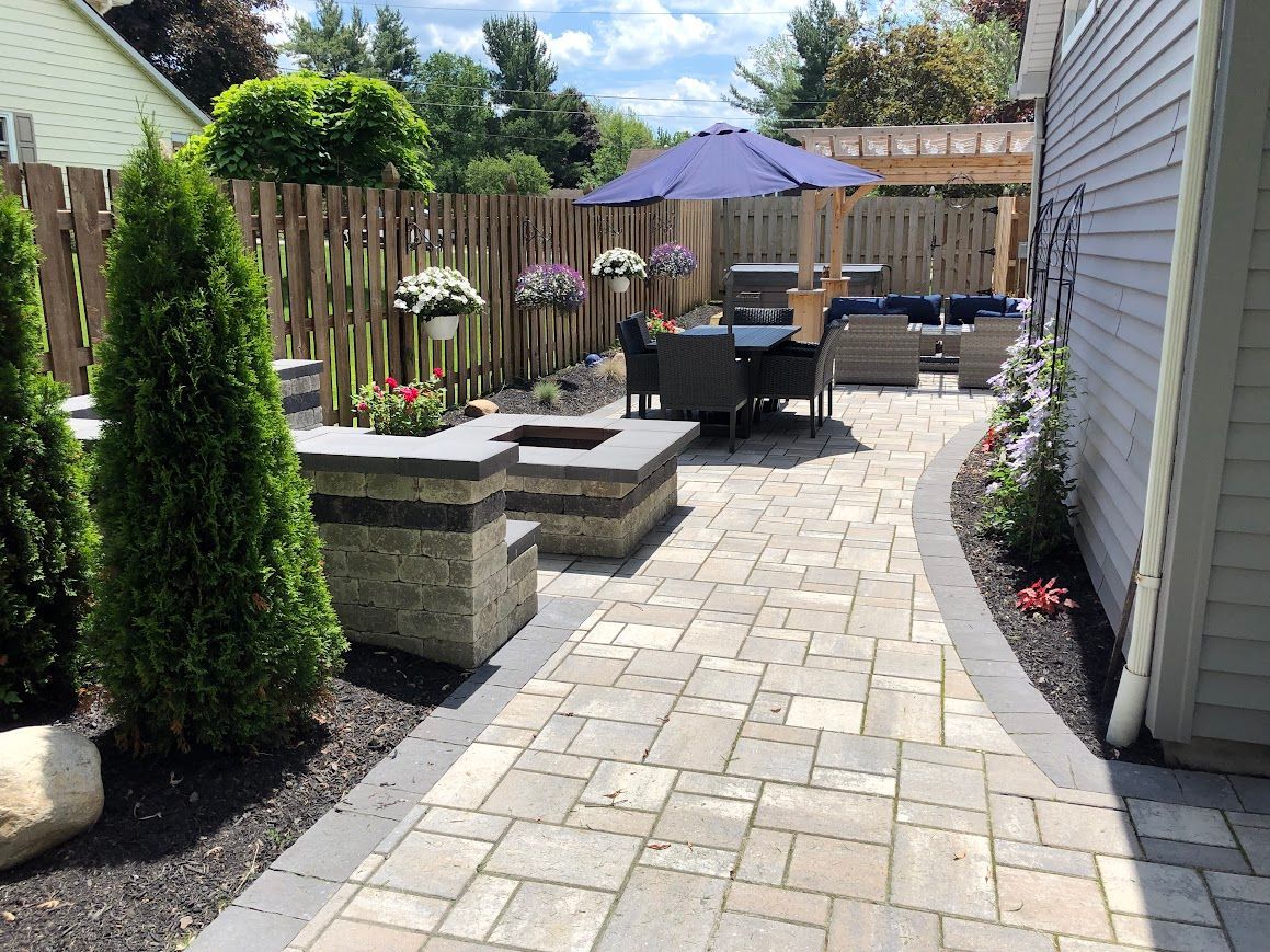 Stone patio with seating area, fire pit, and fence. Umbrella provides shade. Lush greenery surrounds.