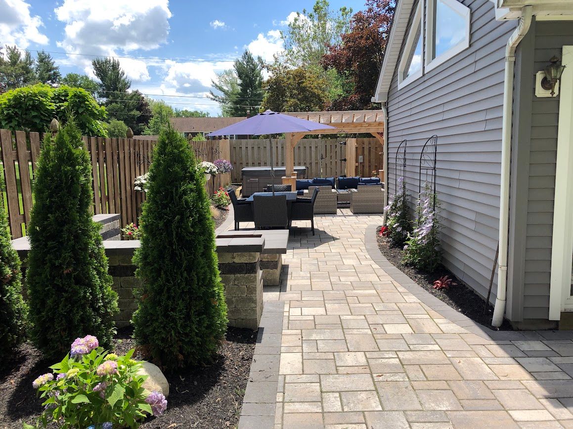 Backyard patio with paved pathway, seating area under umbrella, evergreen trees, and wooden fence.