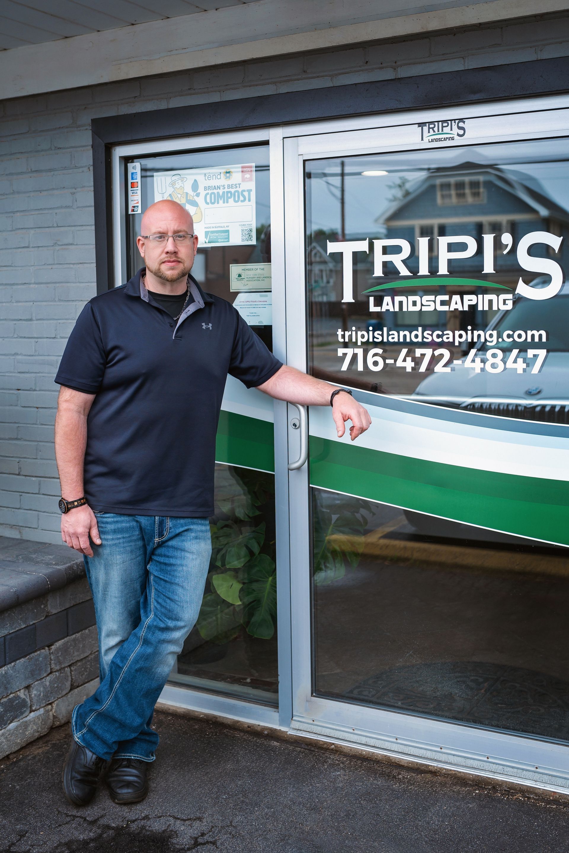 Man leans against the glass door of Tripi's Landscaping office. Black shirt, jeans, and glasses.