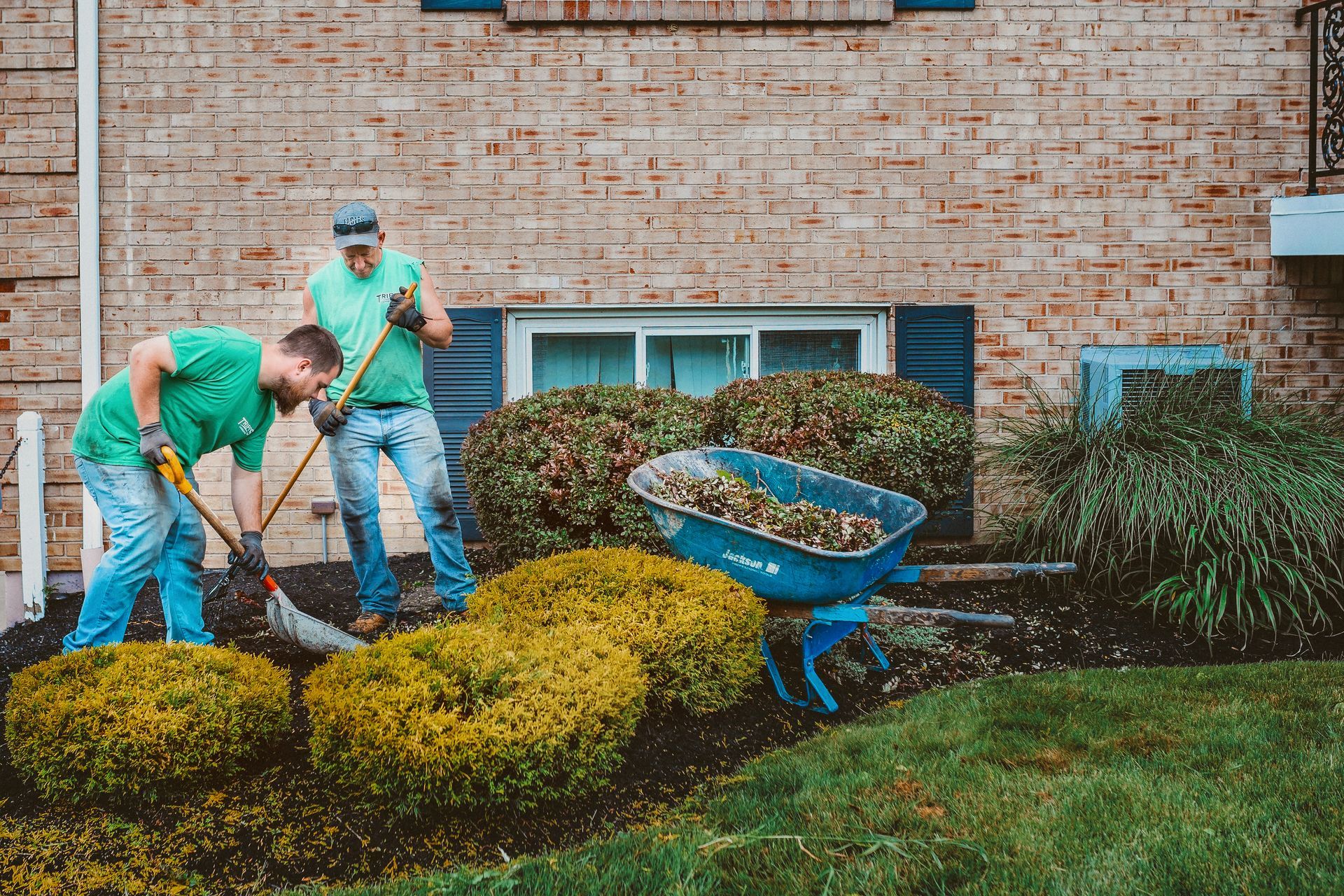 Two workers tending landscaping with tools near a brick building.