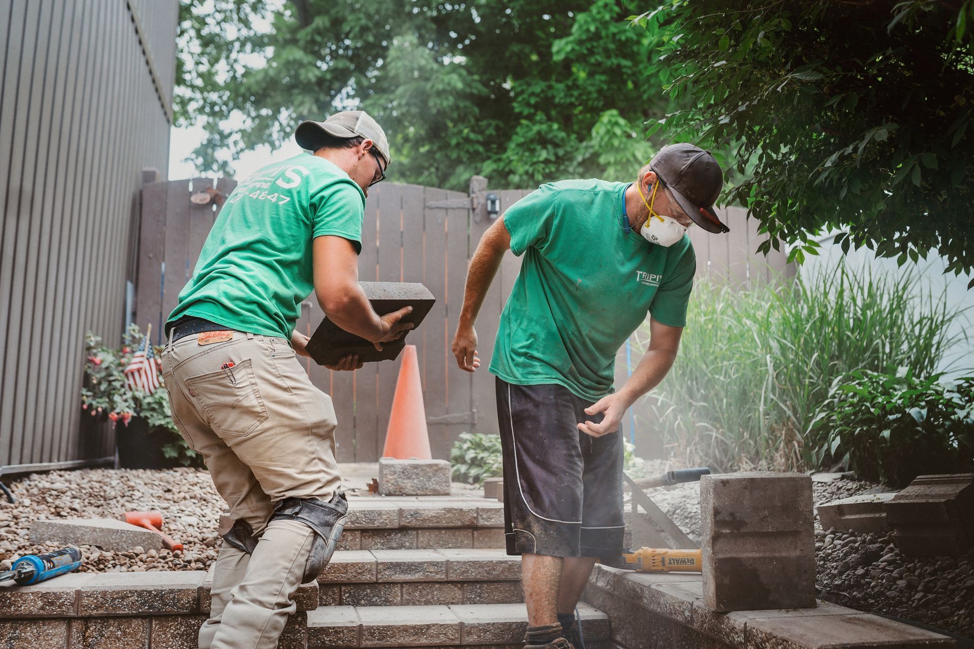 Two workers in green shirts moving stone blocks outdoors. One carries a block, the other brushes dust off the blocks.