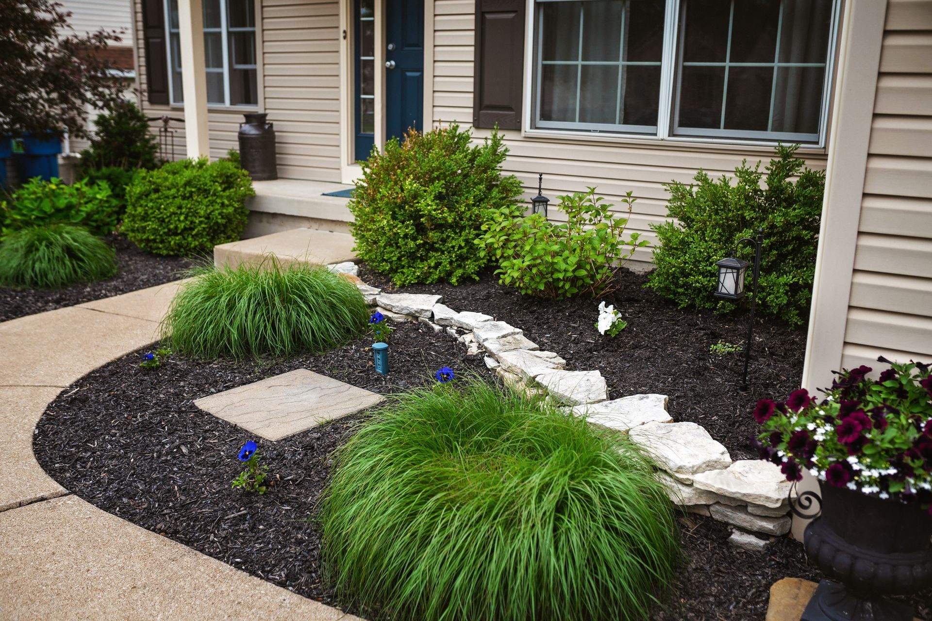 Landscaped front yard with green bushes, ornamental grasses, and black mulch along a curved walkway.