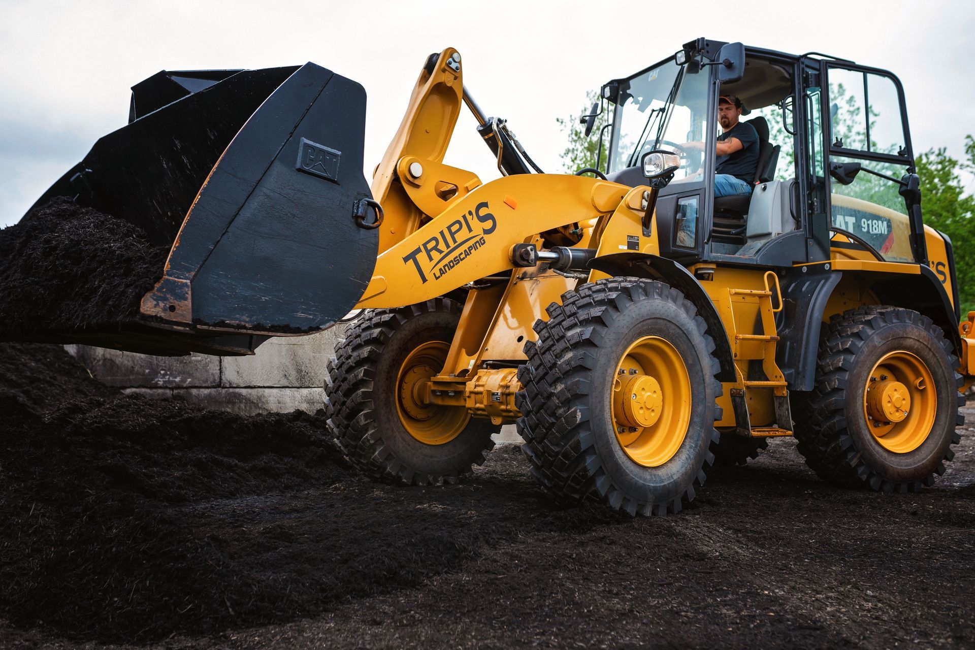 Yellow front-end loader scoops dark material, driver inside. Outdoors, daytime.