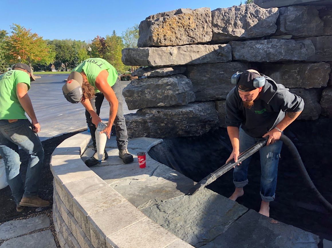 Three people constructing a stone wall and feature, outdoors. One person applying a sealant, and another laying material.