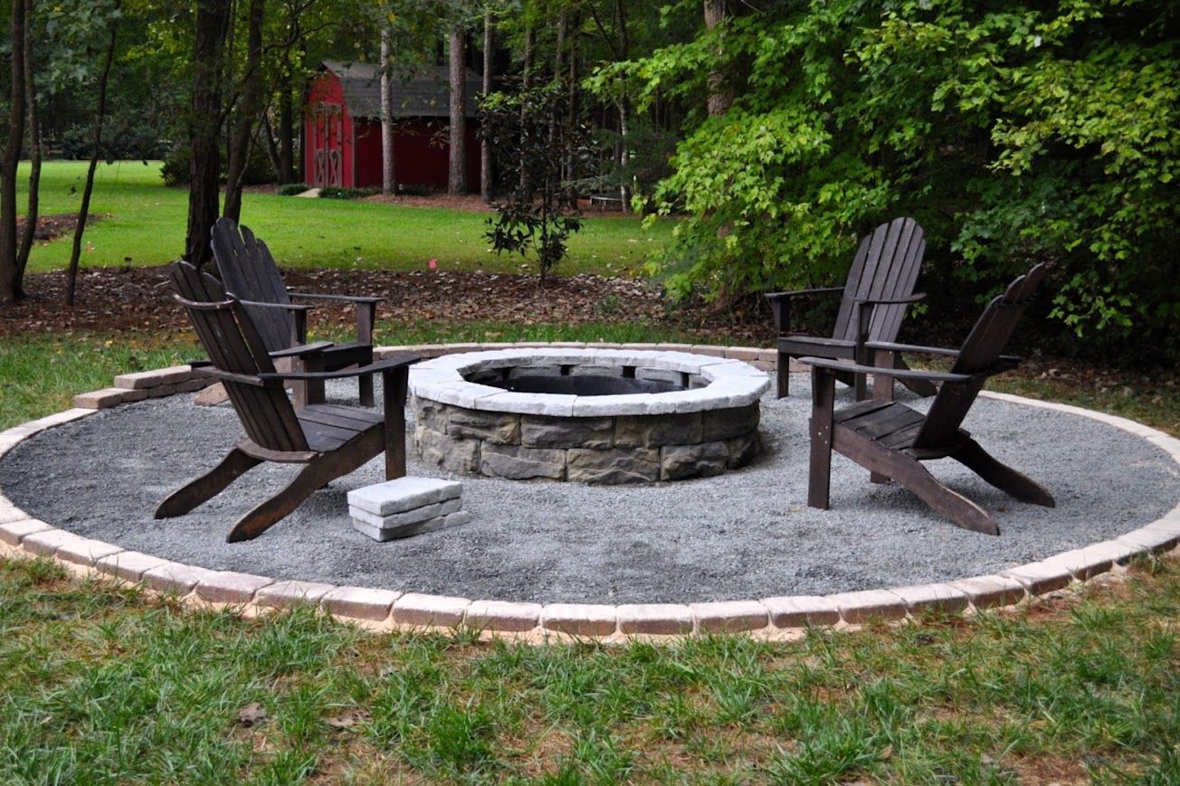 A fire pit with Adirondack chairs on a gravel patio, in a backyard setting.