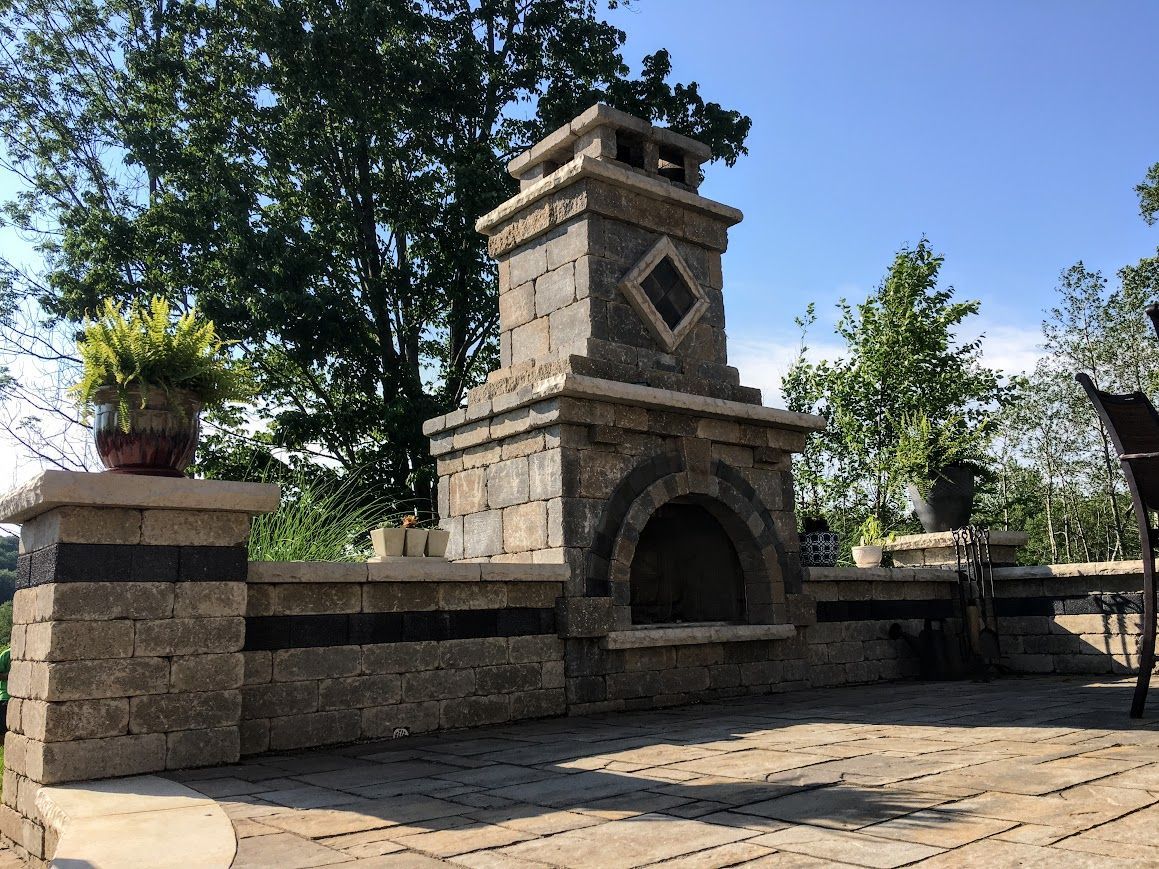 Stone outdoor fireplace with chimney on a patio. Includes potted plants, a blue sky, and surrounding trees.