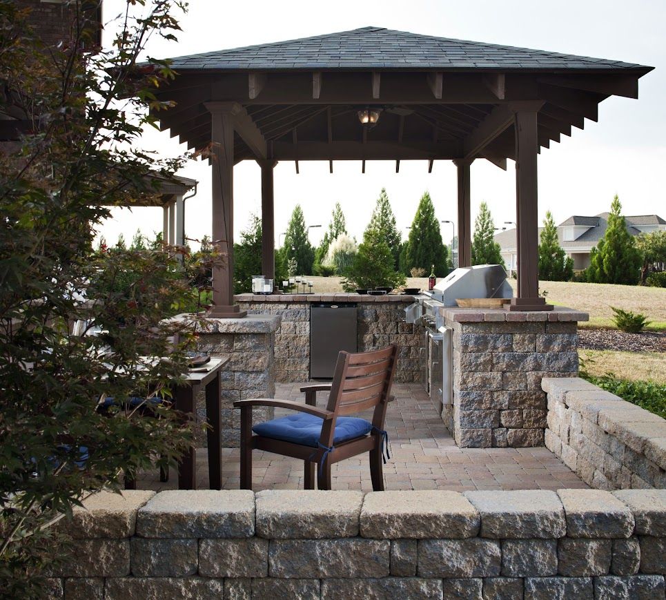 Outdoor kitchen with a gazebo. Stone counters and walls, grilling station. Wooden chair with blue cushion.