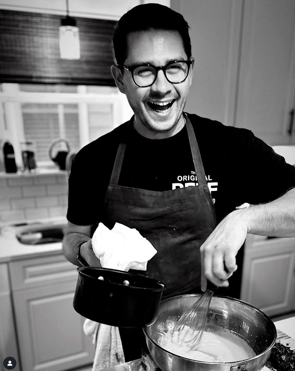 Man in kitchen wearing apron, smiling and whisking batter.