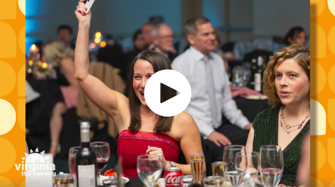 A woman in a red dress raises her hand during an indoor event with guests seated at tables with food and drinks.
