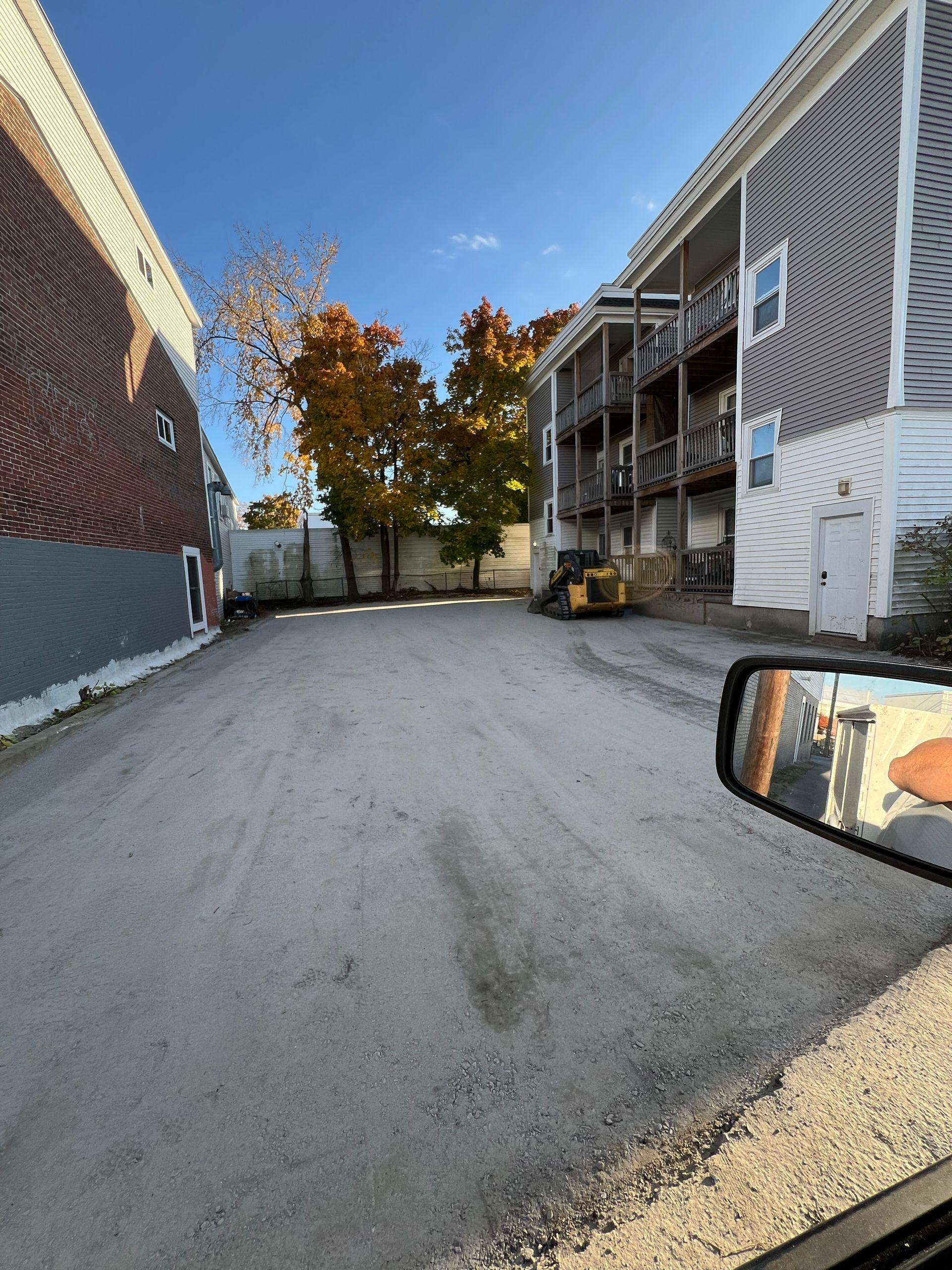Gravel parking area between brick and wood buildings, with a clear blue sky and some fall foliage.