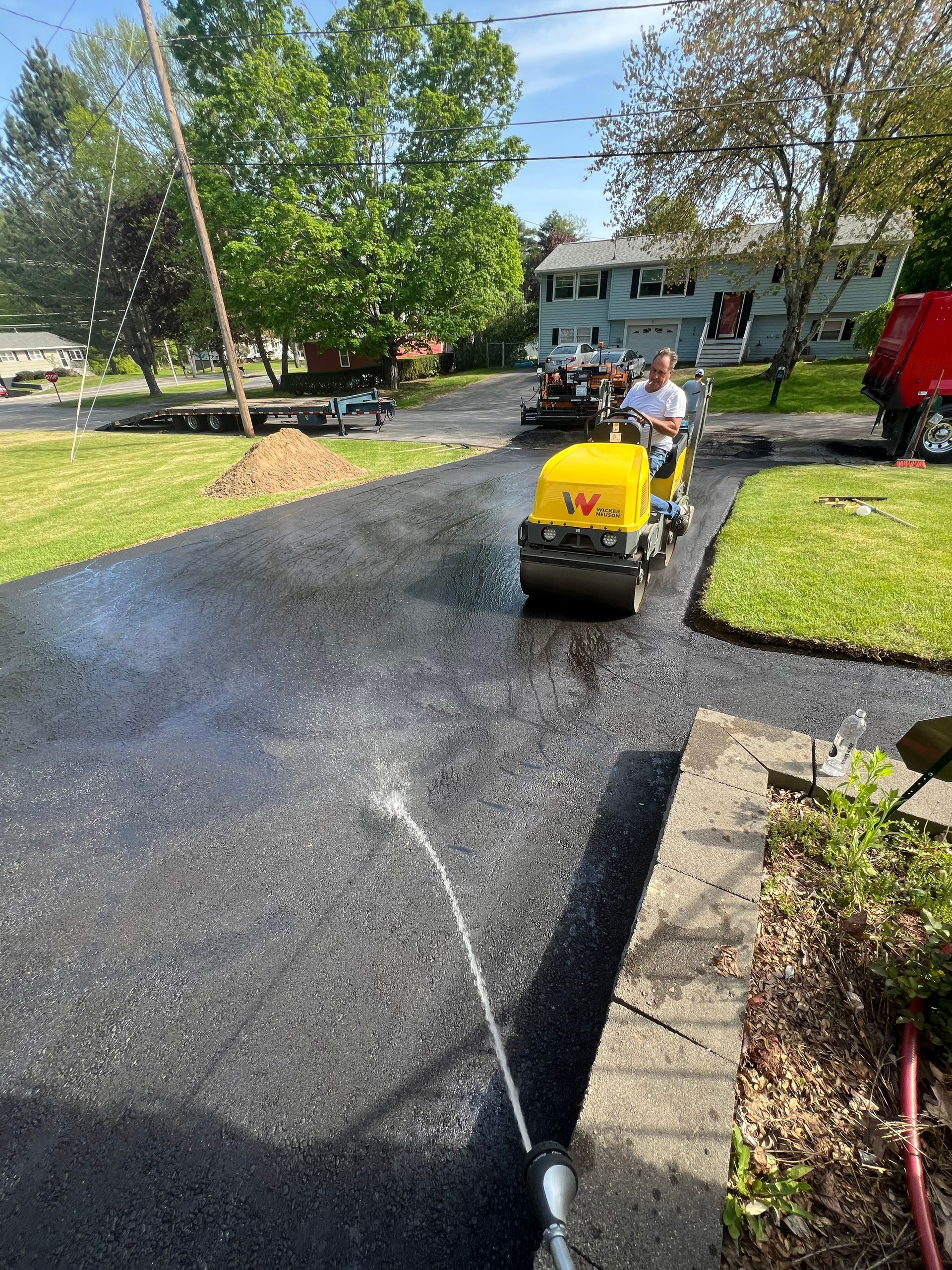 Man operating a yellow asphalt roller on a freshly paved driveway. Houses and trees are in the background.