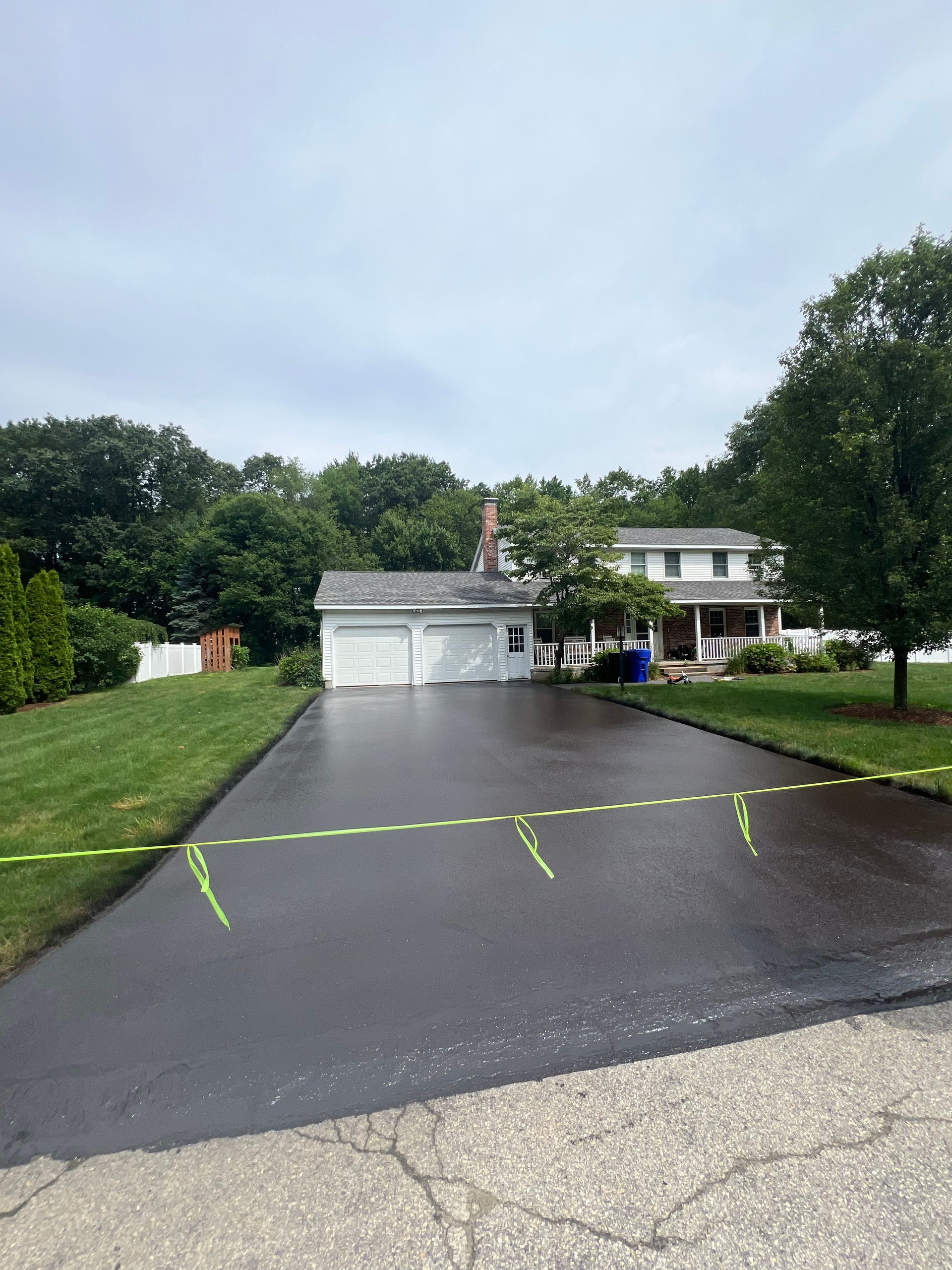 Blacktop driveway leading to a two-car garage and house. Yellow tape crosses the driveway. Overcast sky.