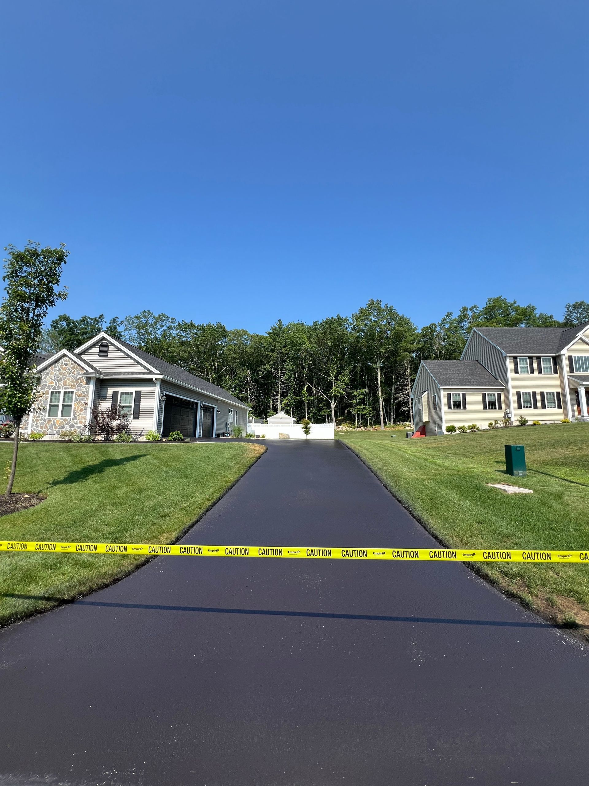 Newly paved asphalt driveway between houses, blocked with caution tape, under a blue sky.