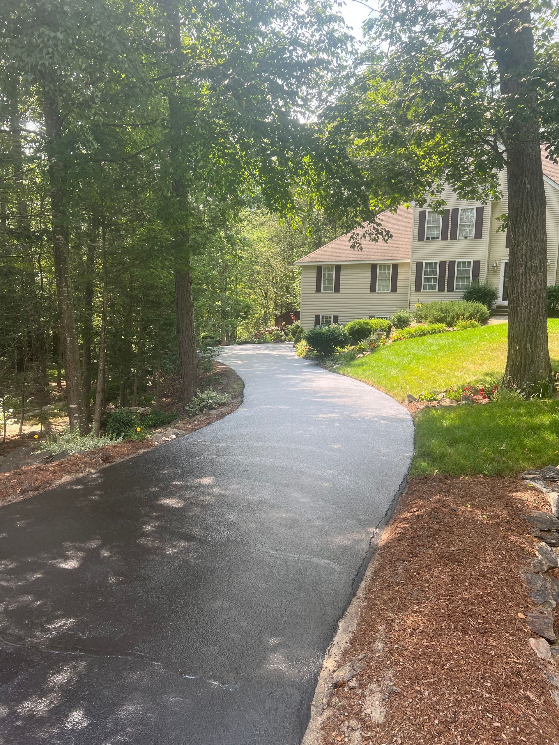 Asphalt driveway winding towards a house with a green lawn, edged with wood chips and trees.