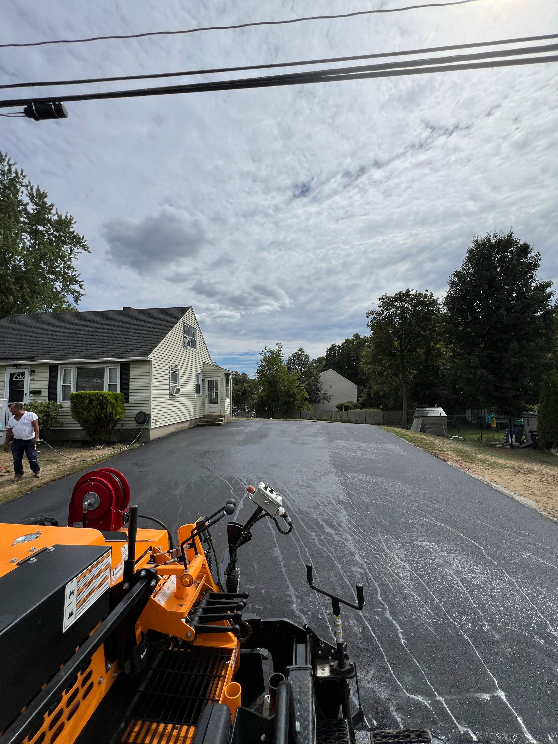Asphalt paving in progress on a residential street; worker, houses, equipment, cloudy sky.