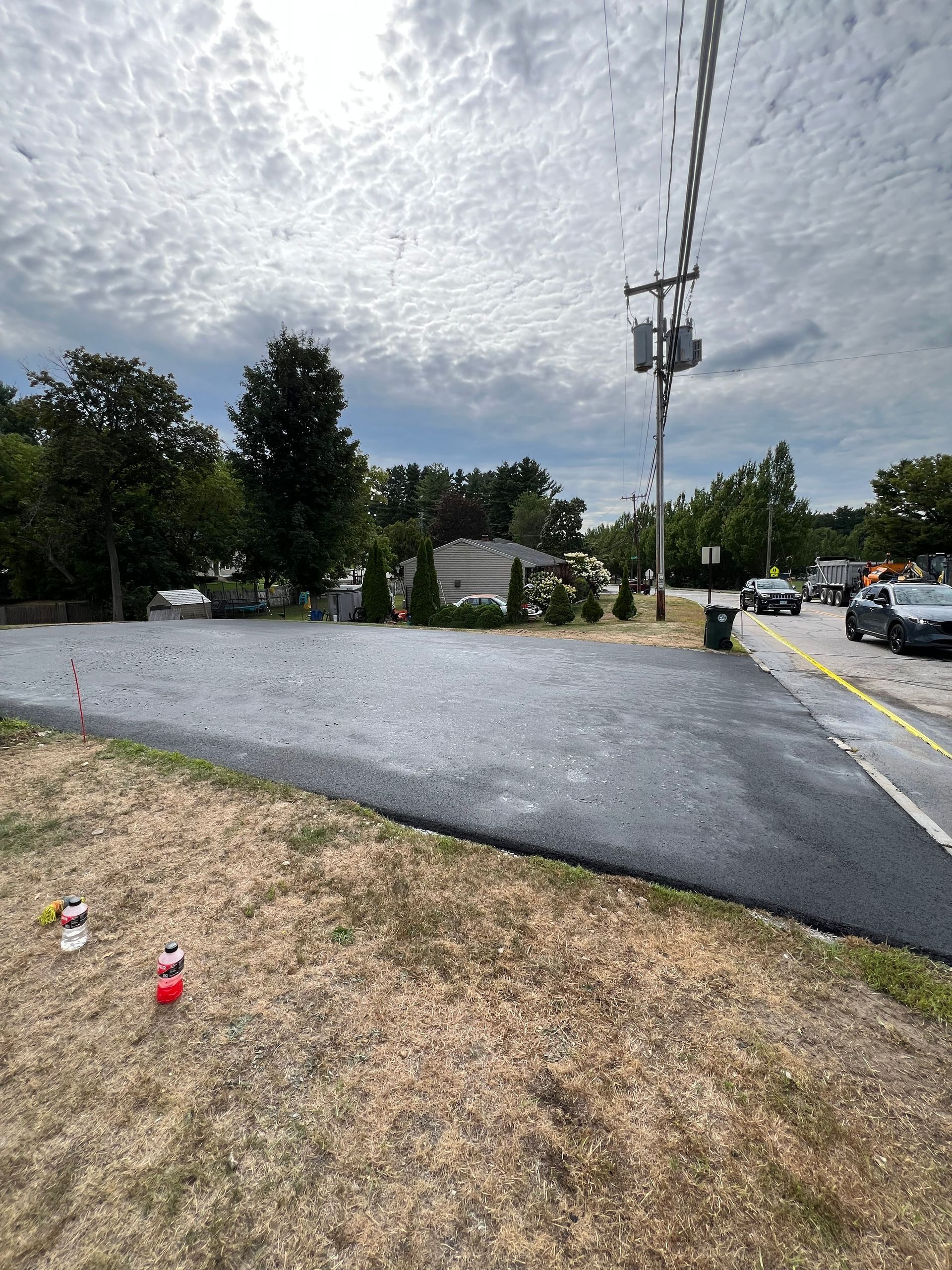 Newly paved asphalt in front of a building, with utility poles, cars, and trees under a cloudy sky.