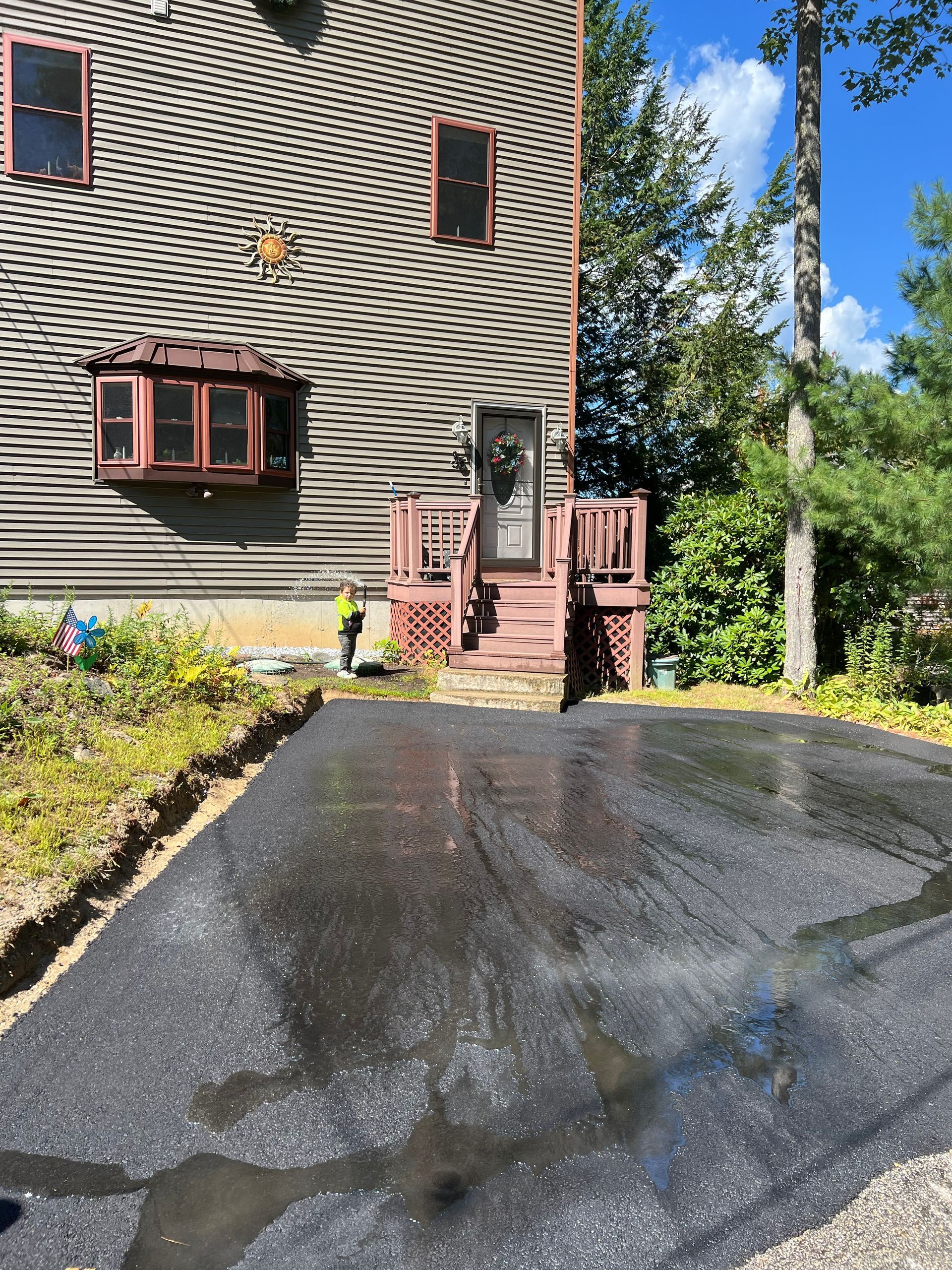 Asphalt driveway leads to a house with a brown roof and a small deck with steps. Sunny day.