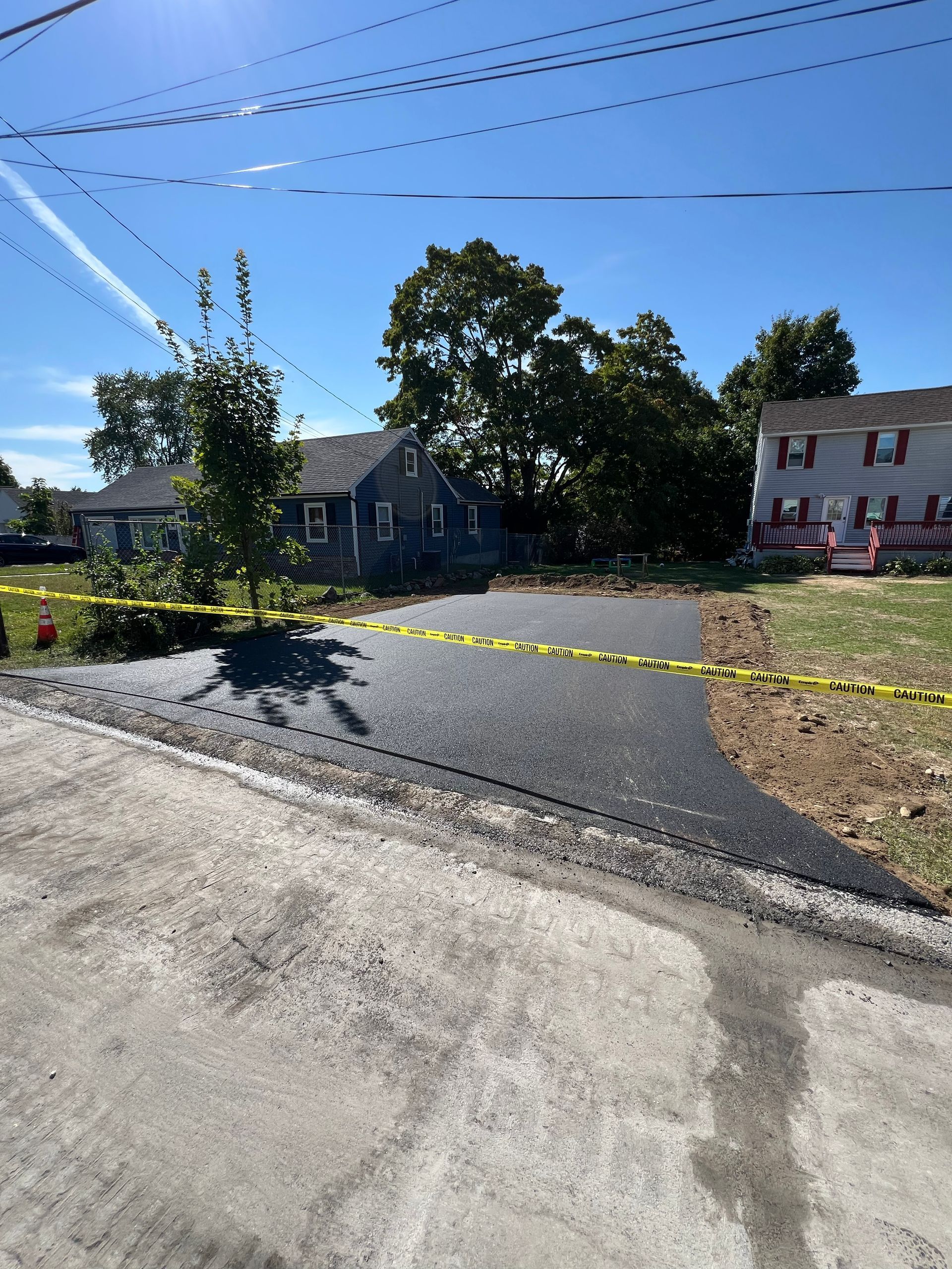 New asphalt driveway cordoned off with yellow tape, houses in the background under a blue sky.