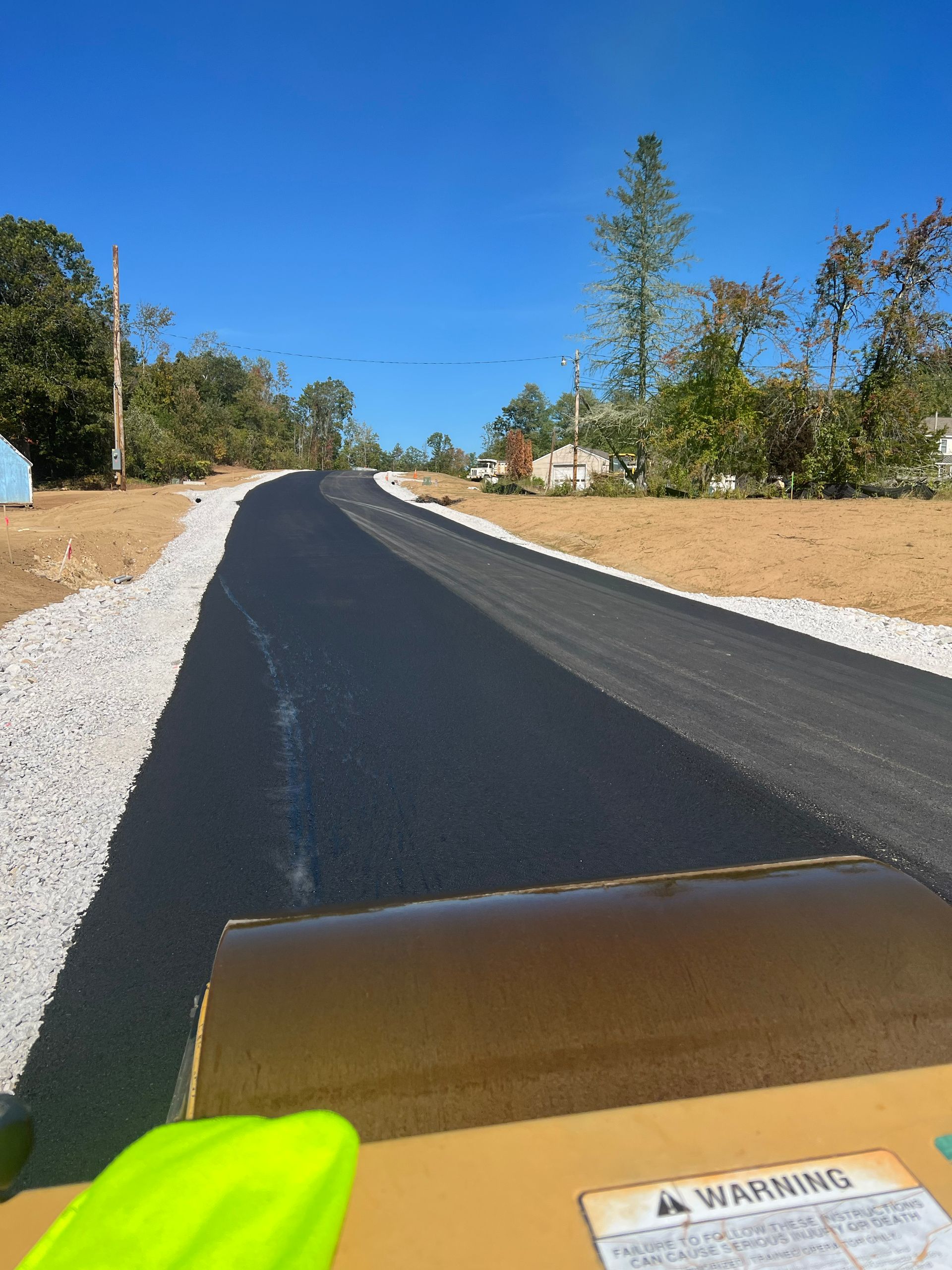 Freshly paved asphalt road, view from roller operator. Gravel shoulders, clear blue sky, trees.