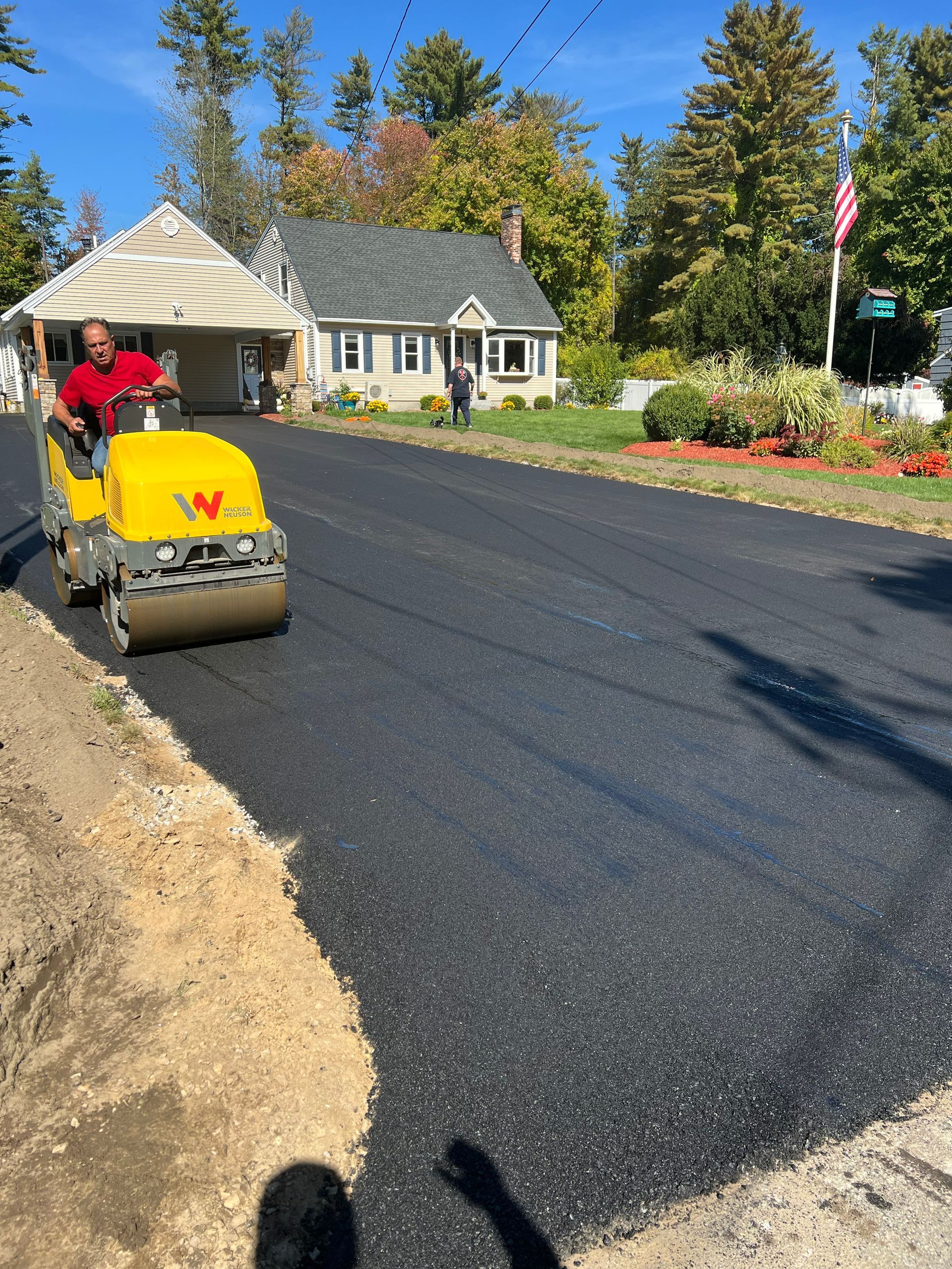Man operating a road roller on fresh asphalt. Houses and trees in background, sunny day.
