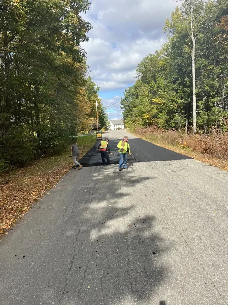 Three workers paving a road surrounded by trees under a cloudy sky.