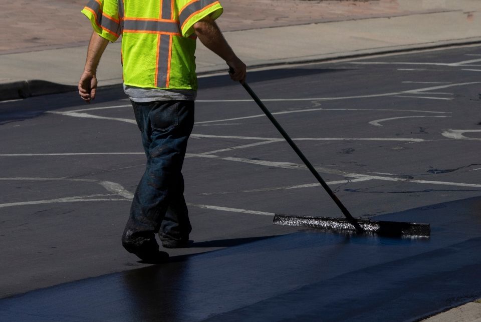Person in reflective vest smoothing fresh asphalt on a road with a long-handled broom.