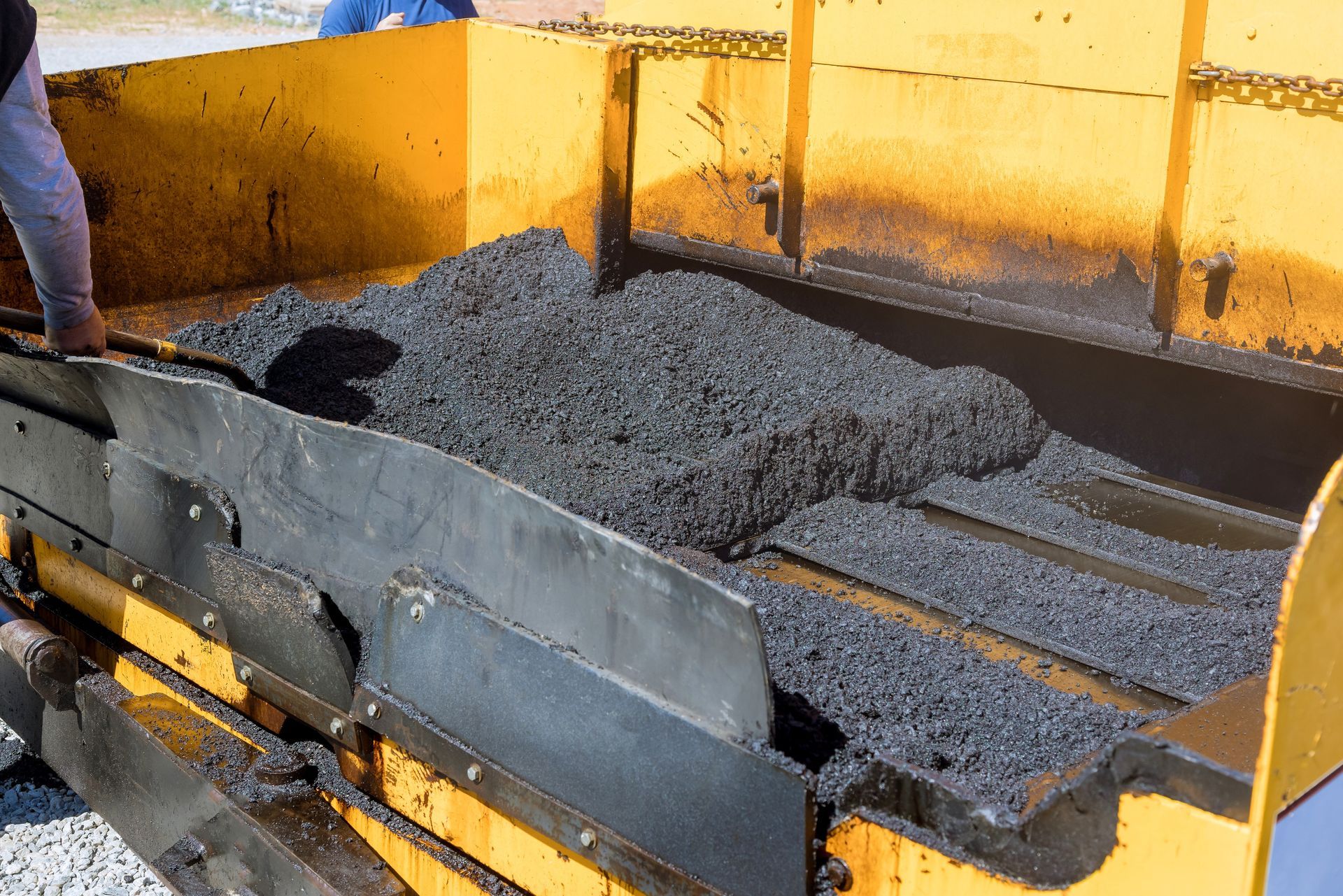 Asphalt being spread from a paving machine, with a worker's arm visible.