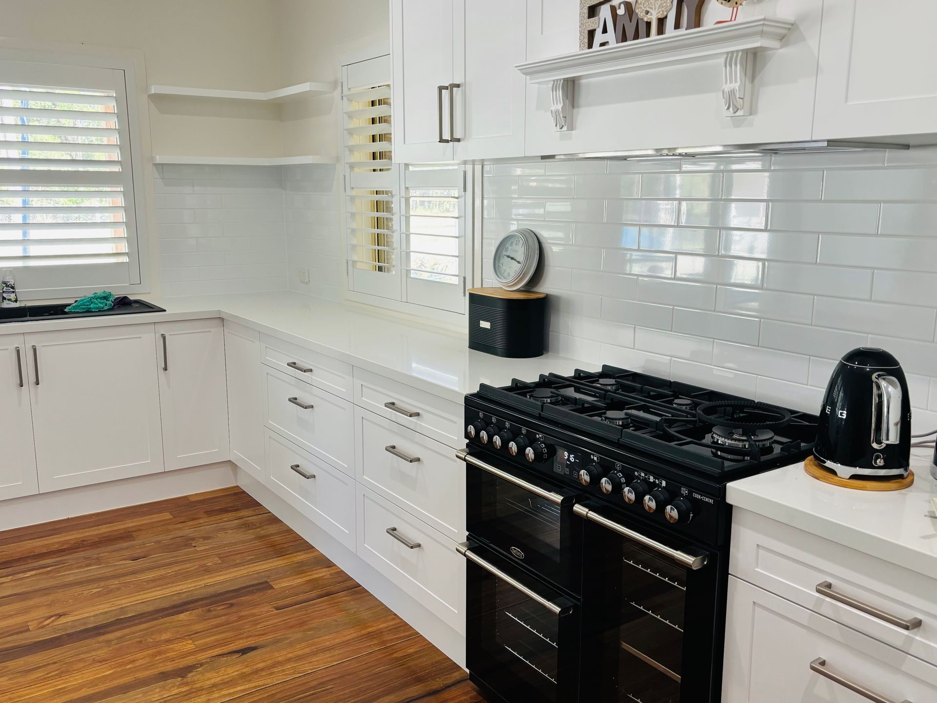White kitchen with wooden floors, black oven, white cabinets, and countertops, shutters on the window.— Piscopo Kitchens Plus In Forster, NSW