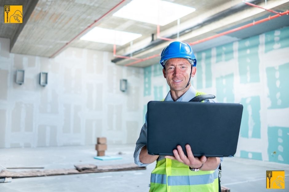 This image shows a drywall contractor standing inside an unfinished building. He is wearing a bright yellow high-visibility safety vest over a light gray shirt, along with a blue hard hat for protection. He holds an open black laptop in his hands, presenting it slightly forward as if showing something on the screen. His posture is confident and engaged. The background of the image reveals an indoor construction site with unfinished walls covered in drywall panels in Richmond, BC