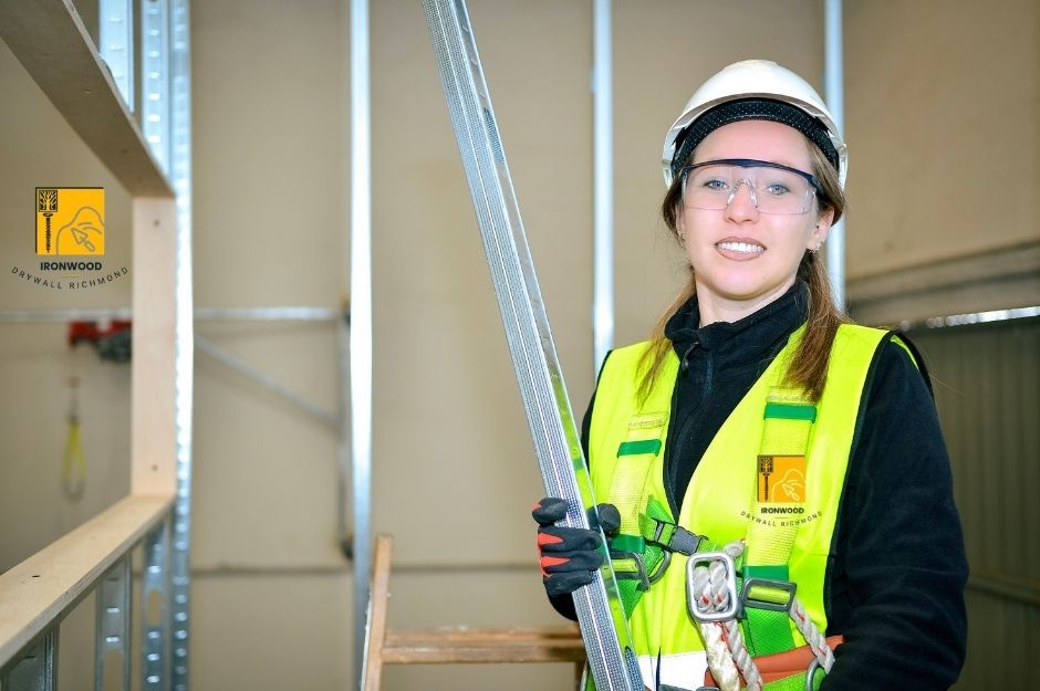 A drywall repair company worker in a bright yellow safety vest and hard hat smiles while holding a measuring tape in a commercial project with metal framing visible on Therrien St and Brunette Ave, Coquitlam, AZ.