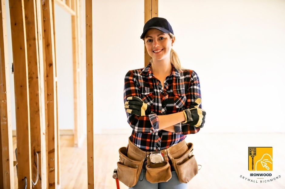Professional female contractor from a drywall repair company wearing plaid shirt, cap, gloves, and a tool belt with arms crossed confidently while overseeing a commercial drywall project. She stands in an unfinished commercial space with exposed wooden framing and partial drywall installation visible. The commercial building renovation is near 190a St and 119B Ave in Pitt Meadows, BC.