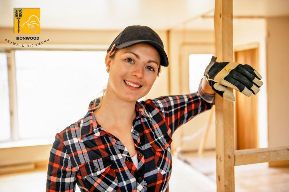 A skilled contractor from a professional drywall repair company stands in a commercial construction site, wearing a black cap and a plaid flannel shirt. She is smiling and leaning on wooden framing with her gloved hands. Behind her, exposed wooden studs and framing are visible. The drywall project is on 32 Ave and Green Ln in Campbell Heights, Surrey, BC.
