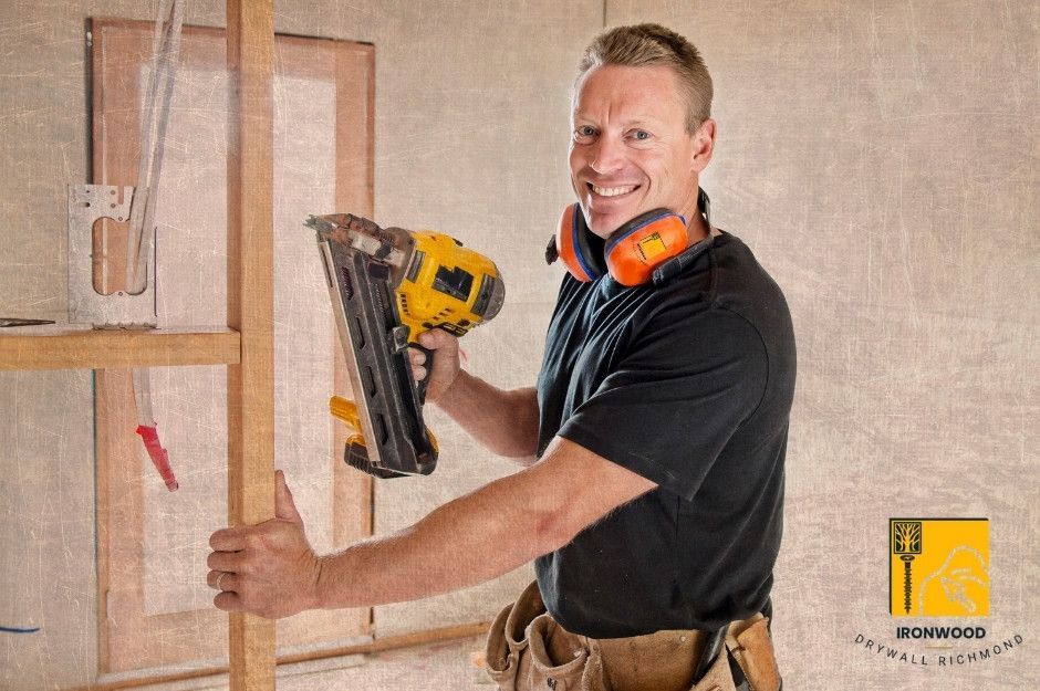 A drywall repair company worker on a commercial project holds a nail gun while installing drywall panels on wooden framing. He wears safety gear including an orange ear protection and a tool belt at Willoughby Way, and 197 St in Willoughby, BC.