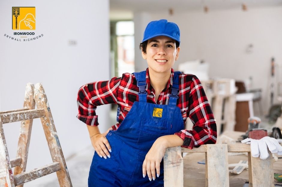 Professional drywall repair contractor wearing blue overalls and plaid shirt, smiling while leaning on wooden ladder in residential construction site with white gyp board. She is working near Delta Ave and Fairlawn Dr, Burnaby, BC.