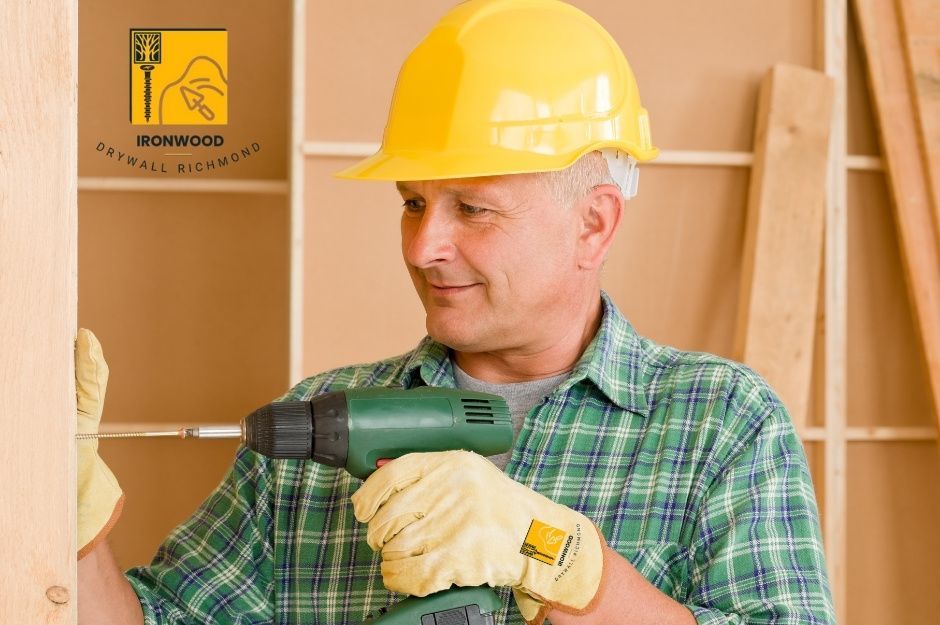 A drywall repair contractor wearing a yellow hard hat and green plaid shirt holds a power drill while working on a residential wall construction. He is positioned next to wooden wall framing with drywall panels visible in the background near 232 St, and 137 Ave in Maple Ridge, BC.
