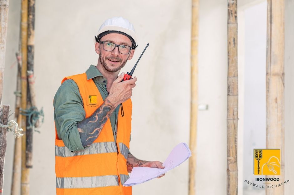 A smiling drywall repair contractor wearing a white hard hat and a safety vest is holding a walkie-talkie at a residential project for wallboard repair near E 9th St, and Ridgeway Ave in Keith-Lynn, North Vancouver, BC.