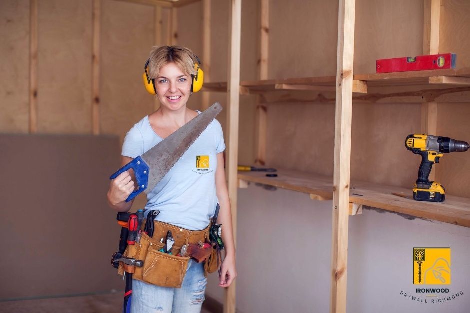 A professional drywall repair contractor stands confidently in a residential construction workspace. Wearing a tool belt around her waist, and is holding a saw. Wooden framing studs are visible along the walls. She is working on a residential drywall project near Eastern Dr, and Audrey Dr, in Mary Hill, Port Coquitlam, BC.