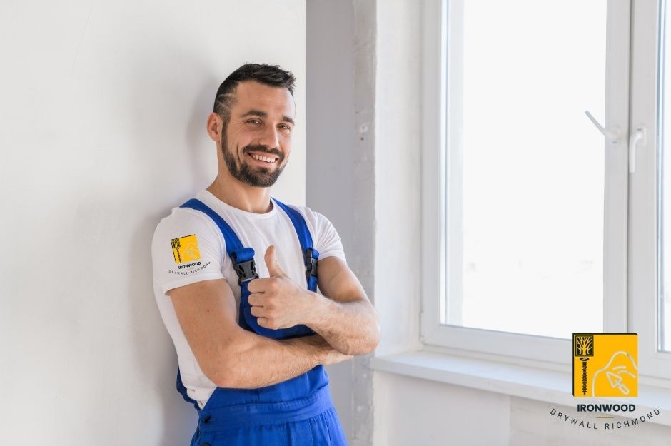 A drywall repair contractor stands confidently in a residential project wearing blue overalls and a white t-shirt. He poses with arms crossed against freshly installed white drywall near W King Edward Ave, and Collingwood St in South Vancouver, BC.