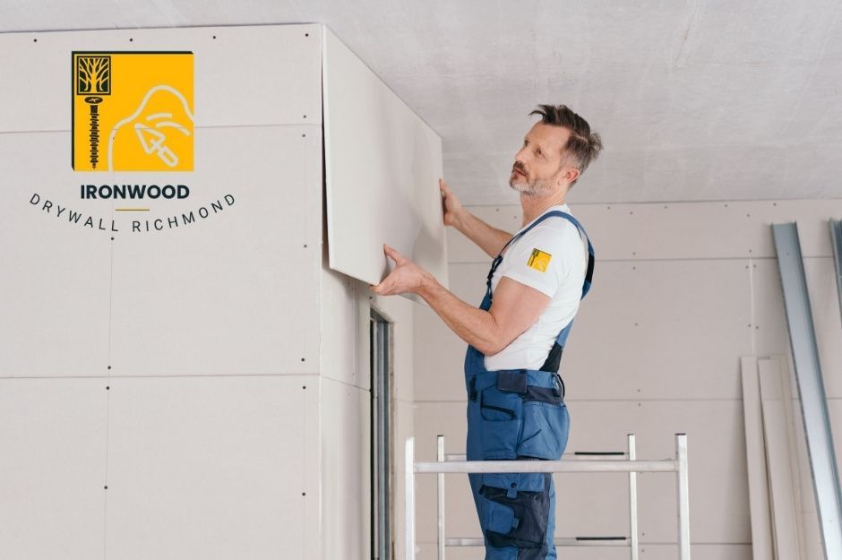 A drywall repair contractor standing on a scaffold carefully installs a drywall sheet to the upper wall corner inside a home. He wears blue work overalls and a white shirt, holding the panel securely in place with both hands. Residential interior under renovation near W 7th Ave, and Collingwood St, Vancouver, BC.