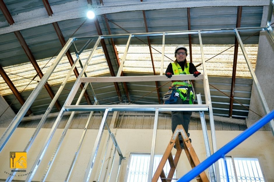 A professional drywall repair contractor is installing metal framing for a commercial drywall system in an industrial building under construction, wearing high-visibility safety gear while working on elevated platform near Everall St, and Thrift Ave in White Rock, BC.
