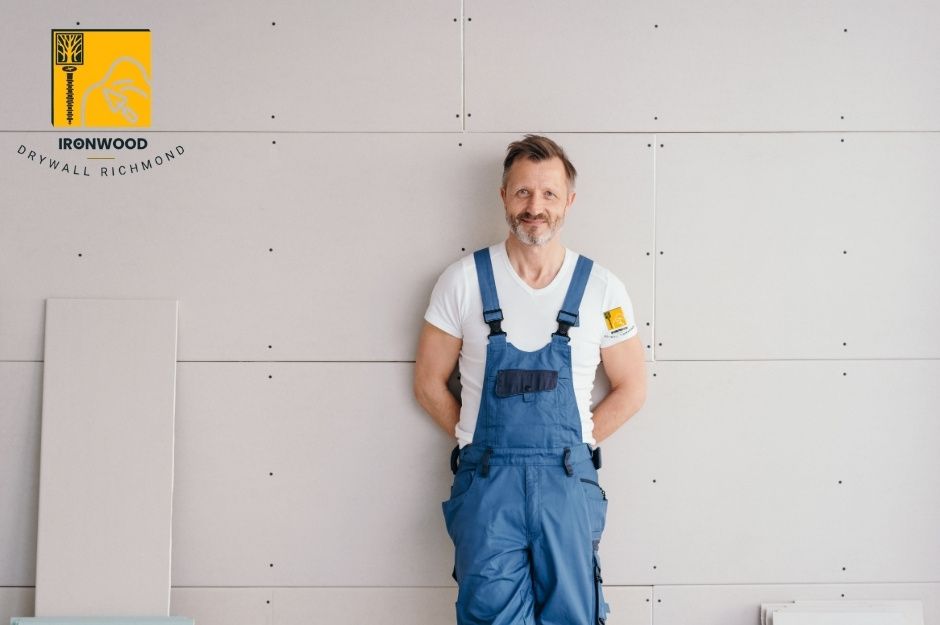 A drywall repair handyman on a commercial project stands against newly installed drywall panels wearing blue overalls and a white shirt. He poses with hands in pockets against the smooth, professionally finished wall surface showing quality craftsmanship in the construction work at 7 Ave, and 56 St in Delta, BC.