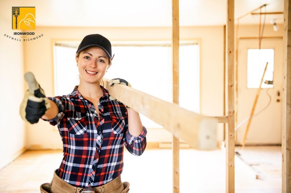 A professional drywall repair handyman in a plaid shirt is holding a wood and giving a thumbs up while working on a residential drywall repair project. She stands confidently in an unfinished room with exposed wooden framing and drywall panels visible in the background. The residential construction is near 199 St and 46a Ave in Langley, BC.