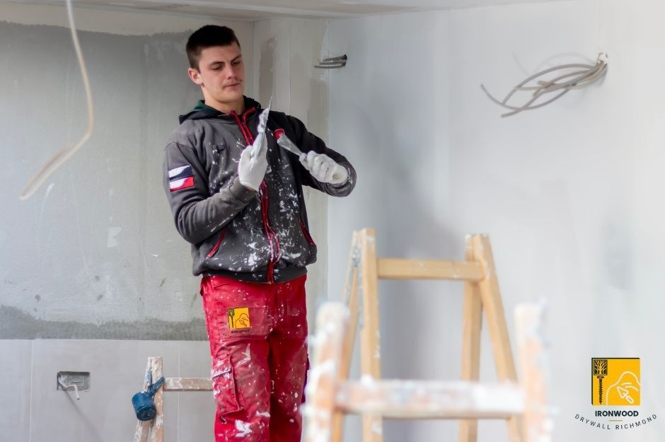 A drywall repair handyman in red pants and a gray jacket holds a sanding tool while standing in a residential room with white walls under construction, with a wooden ladder visible nearby on 7th Ave, and 20th St, New Westminster, AZ.