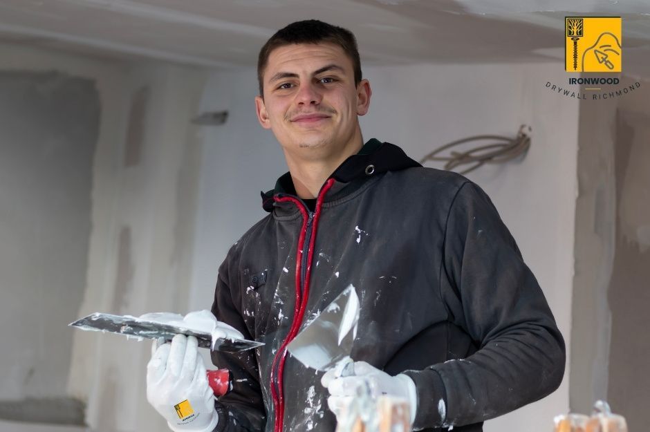 A drywall repair handyman in a dark hoodie holds a putty knife and trowel with white compound in an unfinished residential sheetrock project near Walton Way and Loco Rd, Port Moody, BC.