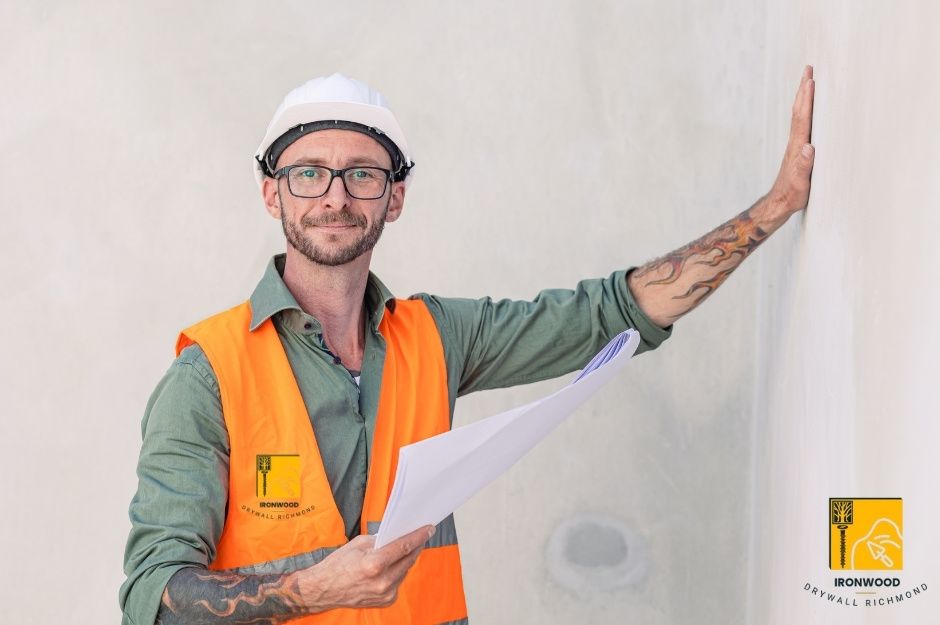 A drywall repair technician in an orange vest and safety glasses stands with a hand against the drywall, inspecting the surface quality of a residential sheetrock project near Pinetree Crescent, and Caulfeild Dr in Caulfeild, West Vancouver, AZ.