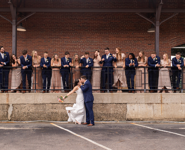 bride and groom kissing while wedding party watches from venue porch